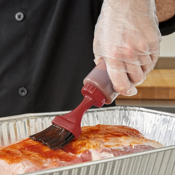 A hand in a plastic glove using a Chef Master basting brush to paint sauce on meat.