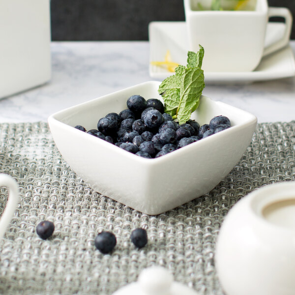 A white square porcelain bowl filled with blueberries with a mint leaf on top.