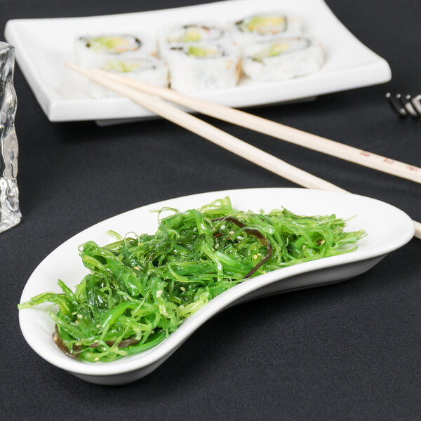 A white Tuxton crescent dish with green seaweed on a table.
