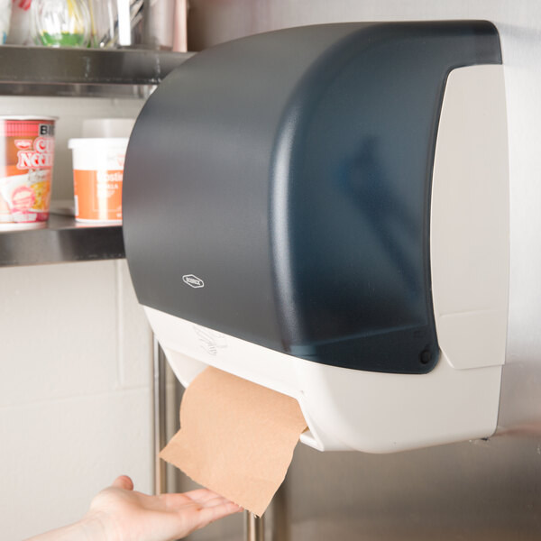 A hand reaching out to a navy blue Bobrick surface-mounted paper towel dispenser with a black plastic cover.