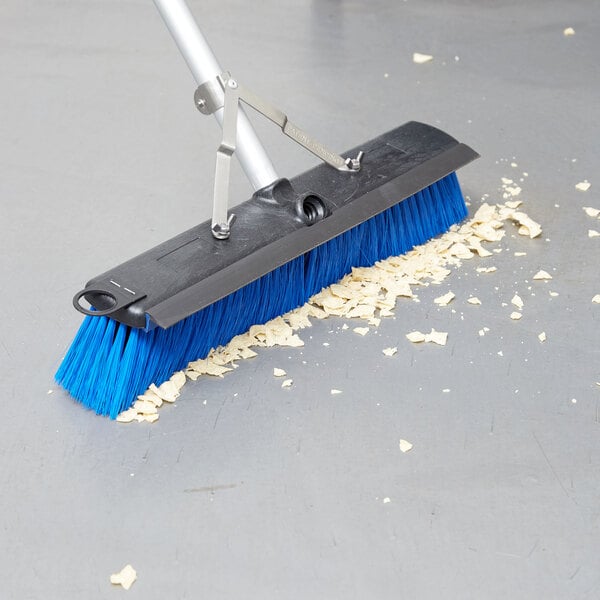 A blue Carlisle commercial push broom with a metal handle and squeegee attachment.