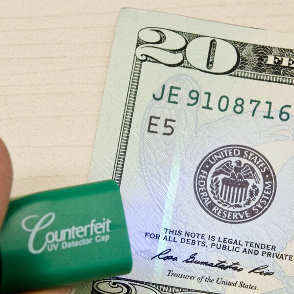 A hand holds a Dri Mark UV light detector pen over a dollar bill.
