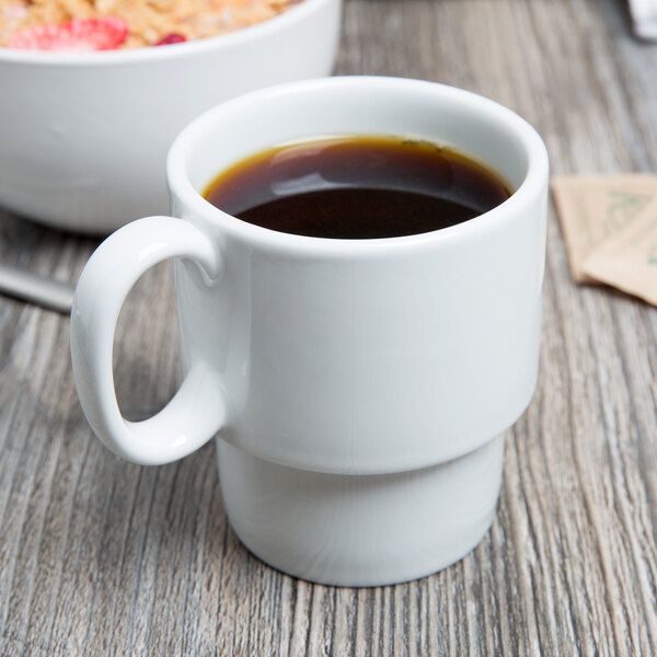 A white Tuxton stackable mug filled with brown liquid next to a bowl of cereal and a cup of coffee.