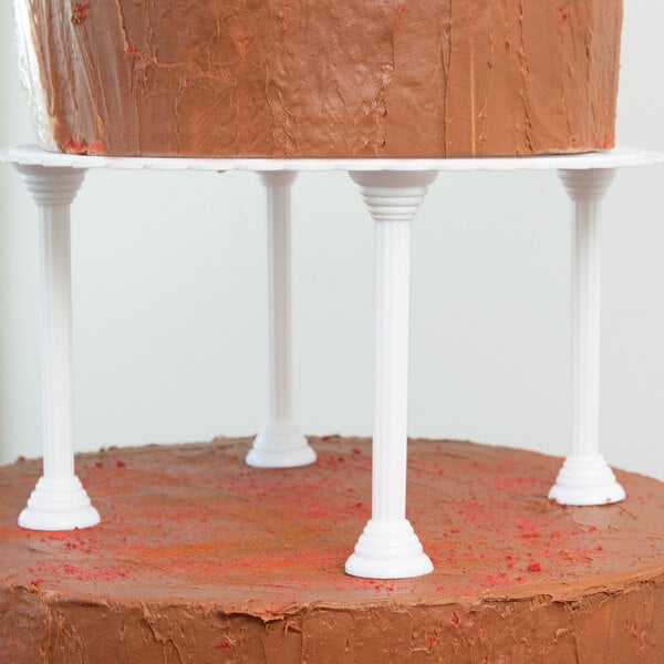 A chocolate cake on a table with a white rectangular Wilton Baker's Best cake stand with 4 white cake pillars.