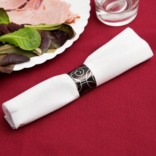 A white linen-like napkin with a silver swirl design wrapped around black plastic cutlery.