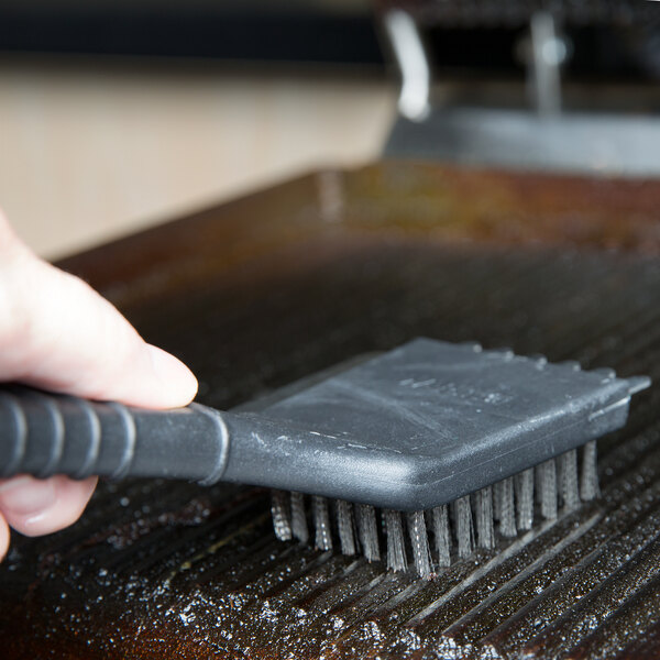 A person cleaning a metal surface on a Waring panini grill with a brush.