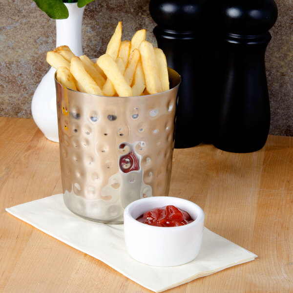 A silver hammered stainless steel cup filled with French fries on a table with a bowl of ketchup.