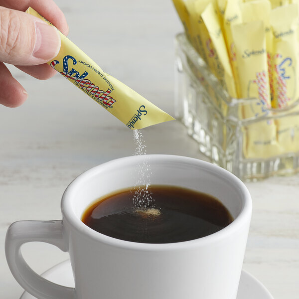 A hand pouring Splenda sugar substitute into a cup of coffee on a table in a café.