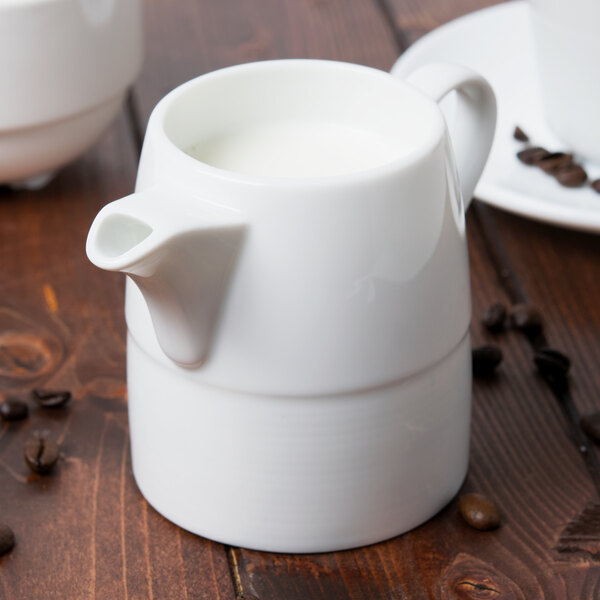 An Arcoroc white creamer with a handle on a white table with coffee beans.