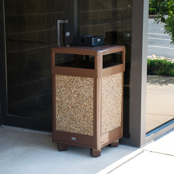 A brown Rubbermaid cigarette and ash receptacle with a hinged top and weather urn sitting outside a building.