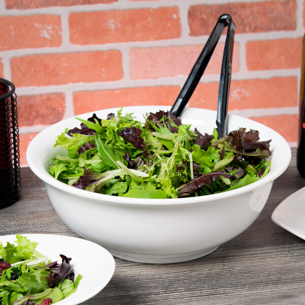 A white porcelain serving bowl filled with green and purple salad on a table with tongs.