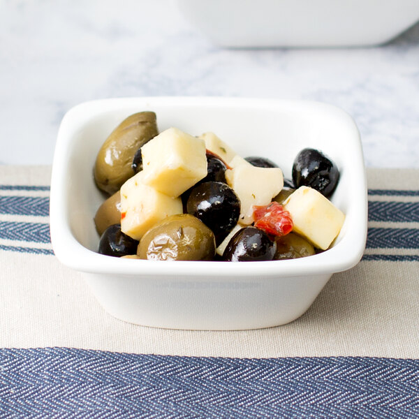 A white square porcelain bowl filled with cheese and olives on a table.