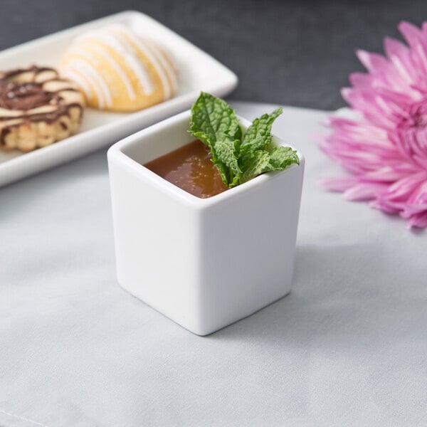 A 10 Strawberry Street white square porcelain bowl with a pastry inside on a table with a cup of liquid and mint leaves.