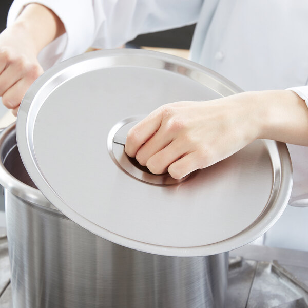 A person in a white chef's uniform holding a Vollrath stainless steel pot lid over a pot.