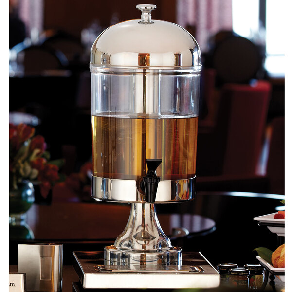 An American Metalcraft stainless steel beverage dispenser on a table with a glass container of liquid in it.