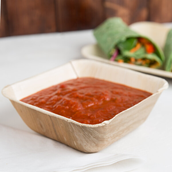 A white table with a bowl of red sauce in a brown Eco-gecko palm leaf bowl and green vegetable wraps.