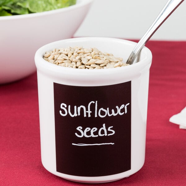 A white Cal-Mil melamine crock filled with sunflower seeds with a spoon in it.