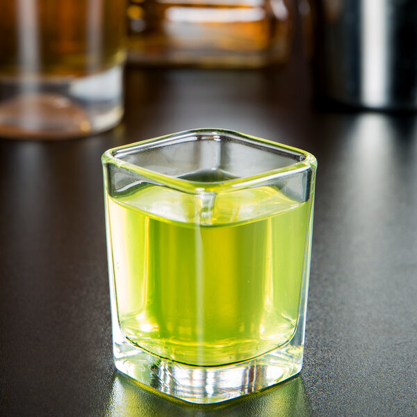An Arcoroc square shot glass with green liquid in it on a table.