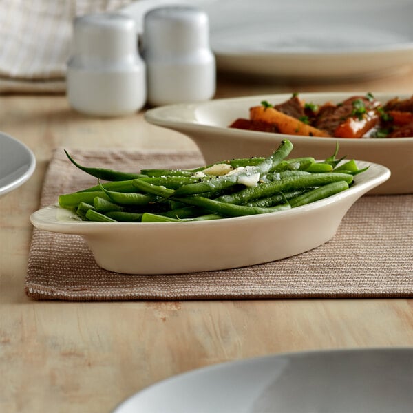 An Acopa ivory oval stoneware rarebit dish filled with green beans on a table with a plate of food.