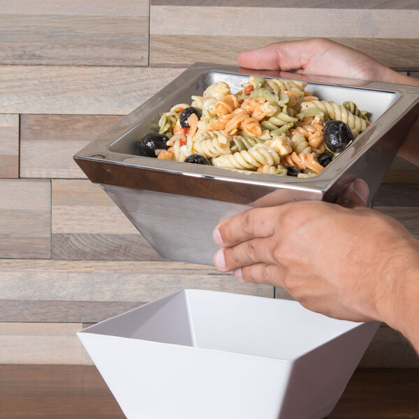 A hand holding a Cal-Mil stainless steel bowl of pasta with olives.
