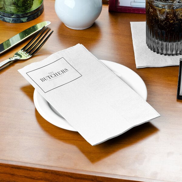A white customizable dinner napkin on a plate on a table.