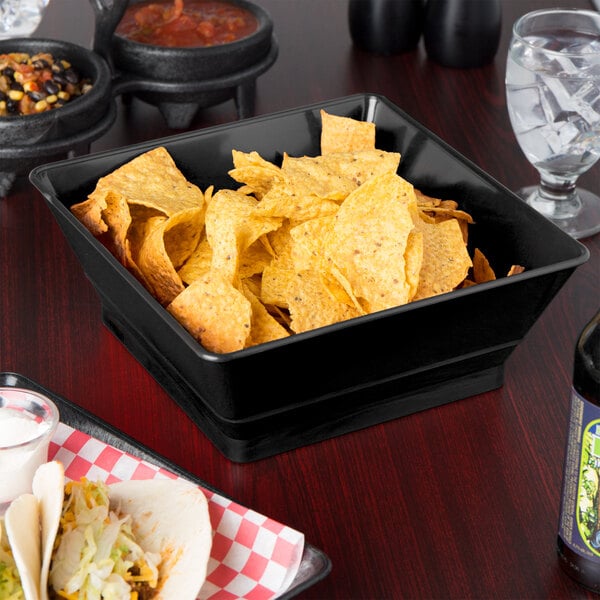 A black square serving basket filled with chips on a table with drinks.