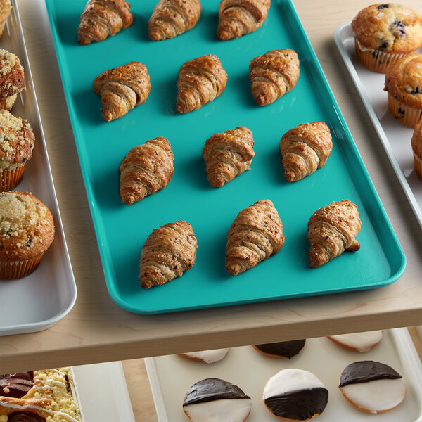 A green Cambro market tray of croissants and muffins on a bakery display counter.