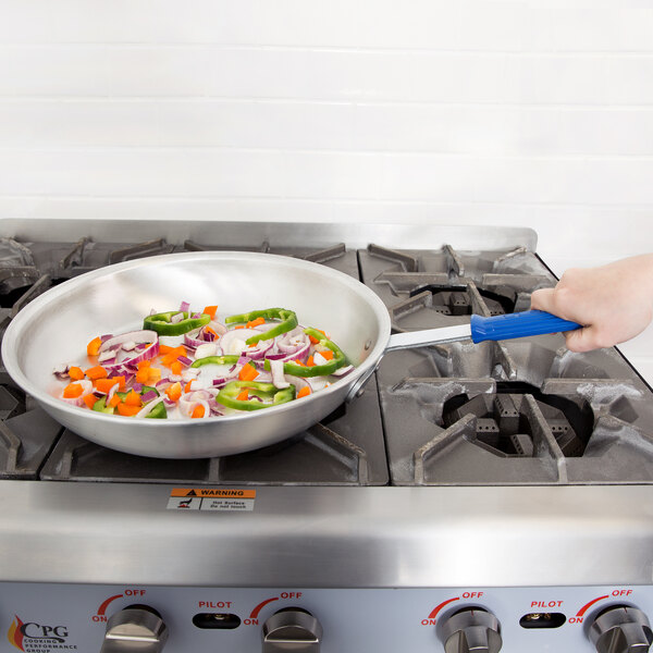 A hand with a blue Vollrath Wear-Ever pan cooking vegetables on a stove.