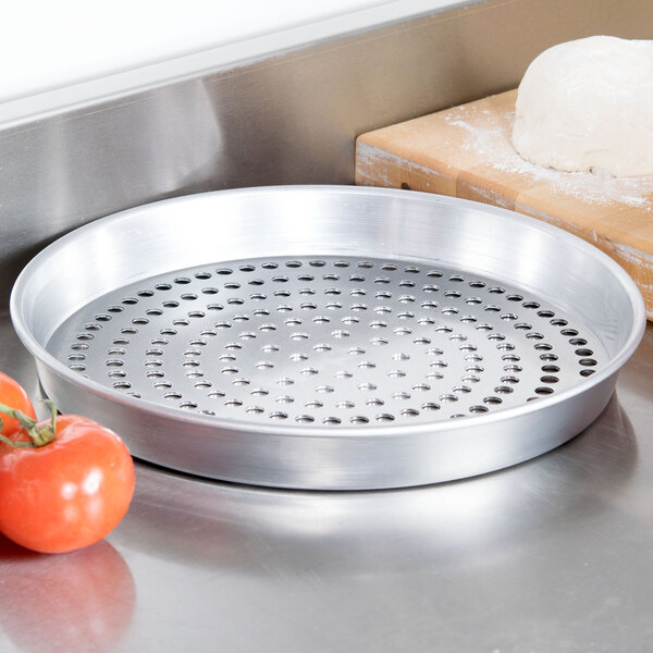 An American Metalcraft heavy weight aluminum pizza pan with perforated holes on a table next to tomatoes.