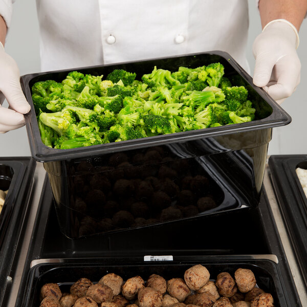 A person in white gloves holding a black Cambro plastic food pan filled with broccoli.