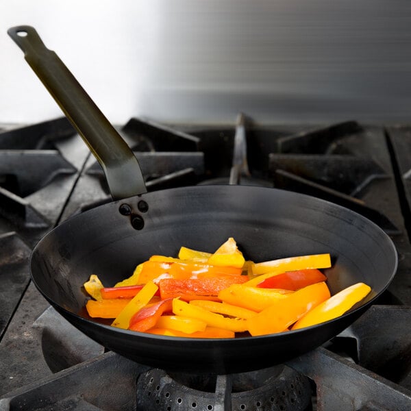 A Vollrath French style carbon steel fry pan with food cooking in it on a stove.