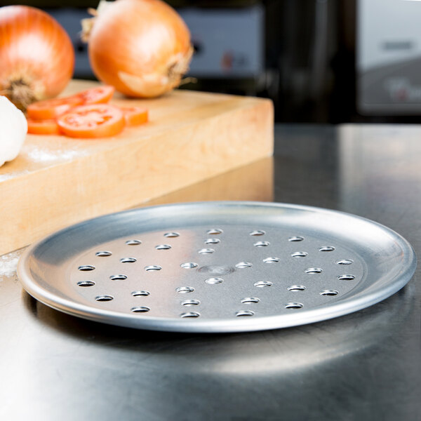 An American Metalcraft perforated aluminum pizza pan on a white background next to onions and garlic.