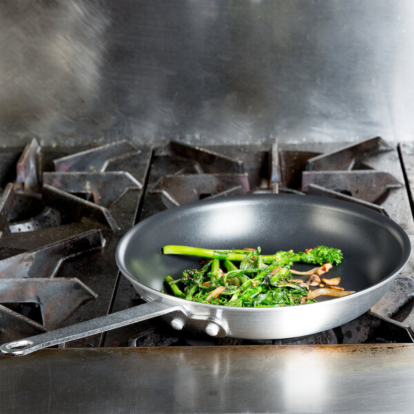 A Vollrath Arkadia non-stick fry pan with broccoli and mushrooms on a stove.