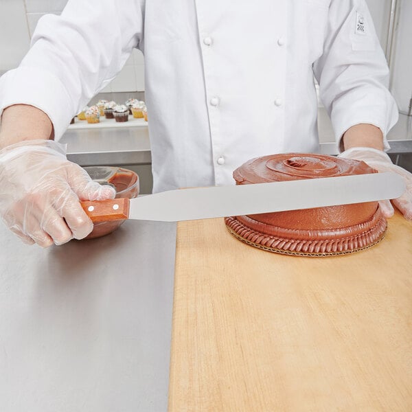 A person in a white coat using a Thunder Group straight baking spatula with a wood handle to cut a brown cake.