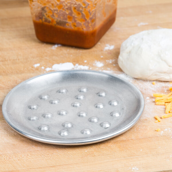 An American Metalcraft aluminum pizza pan with pizza dough on a table.