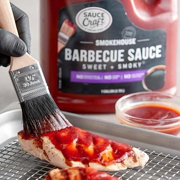 A hand using a paint brush to spread Sauce Craft Sweet and Smoky BBQ Sauce on food on a grill.