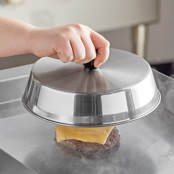 A hand holding an American Metalcraft stainless steel basting cover over a cheeseburger on a grill.