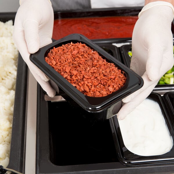 A person in gloves holding a Cambro black plastic food pan over food on a counter.