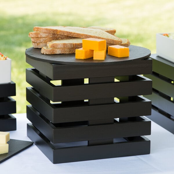A black wooden square crate riser with black and white containers of food on it.