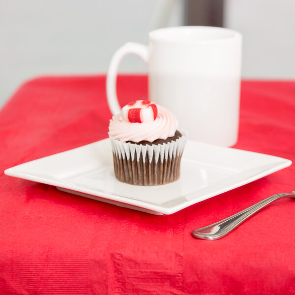 A cupcake with a peppermint candy on top sits on a white Arcoroc square porcelain plate next to a white mug.