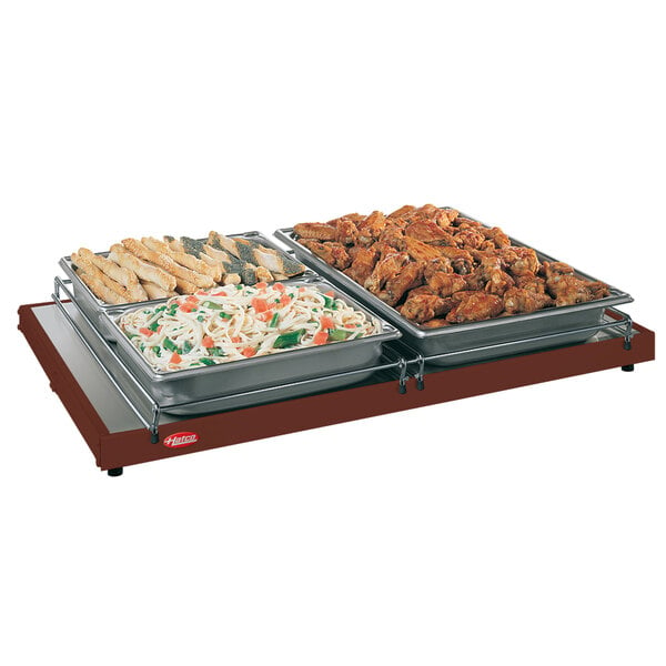 A Hatco heated shelf holding trays of pasta, bread sticks, and chicken wings on a table.