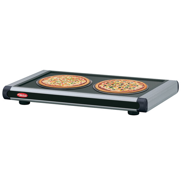 Two pizzas on black and silver trays on a Hatco heated shelf.