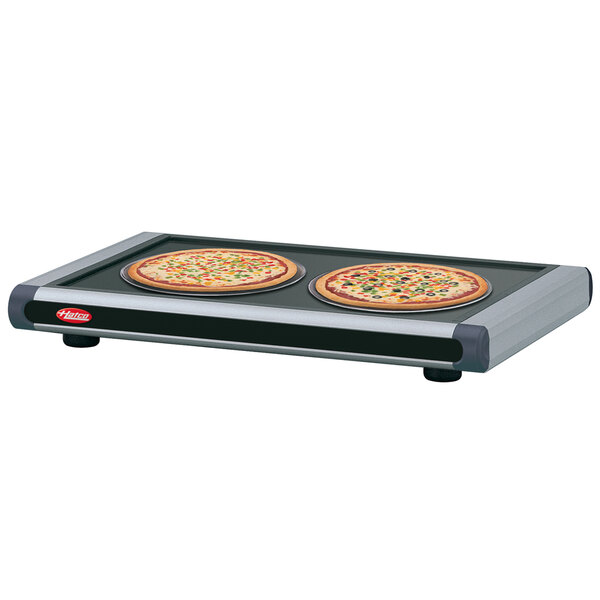 Two pizzas on a table with a Hatco heated shelf with dark gray and black trays.
