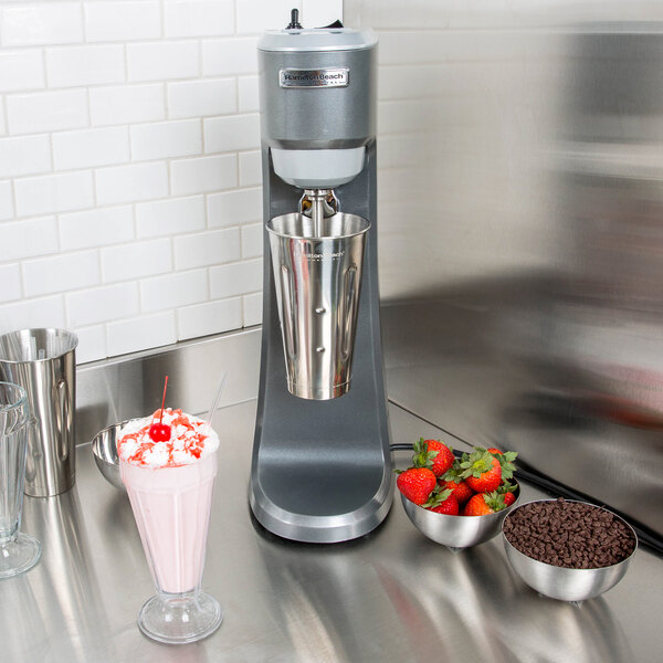 A Hamilton Beach drink mixer with a glass of strawberry milkshake on the side and bowls of strawberries and chocolate chips.