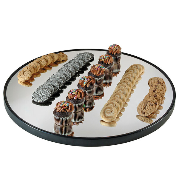 A Cal-Mil round mirror tray with a raised black rim holding cookies and cupcakes on a table in a bakery display.