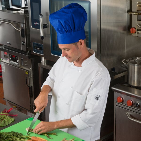 A man wearing a Choice royal blue chef hat and uniform cutting vegetables on a cutting board.