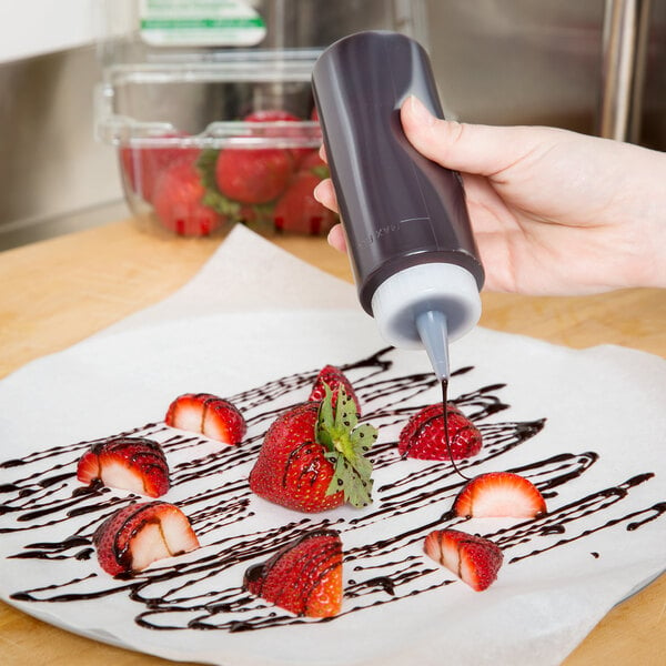 A person using a Wilton squeeze bottle to pour chocolate syrup onto strawberries on a white paper.