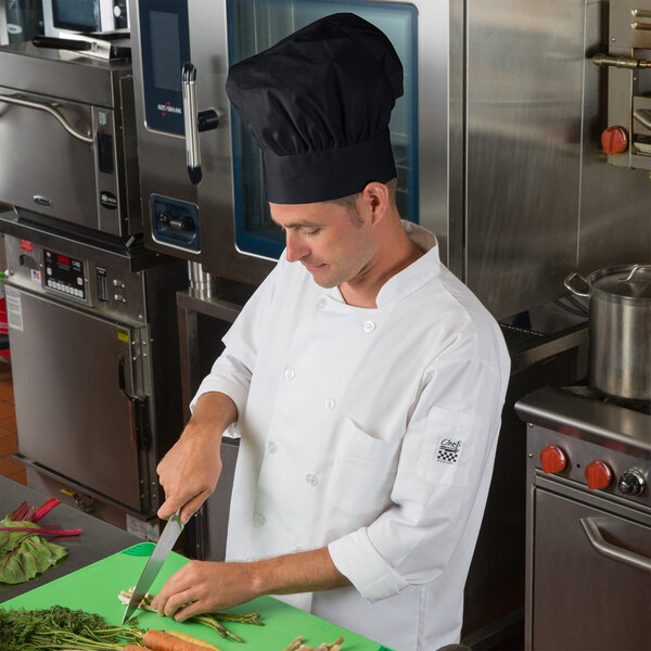 A man in a Choice black customizable chef hat and uniform cutting vegetables on a cutting board.