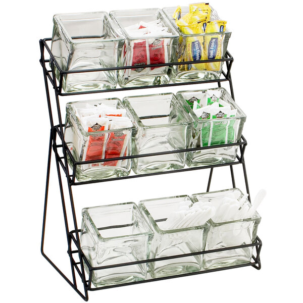 A black iron Cal-Mil three tier rack holding glass jars filled with condiments.