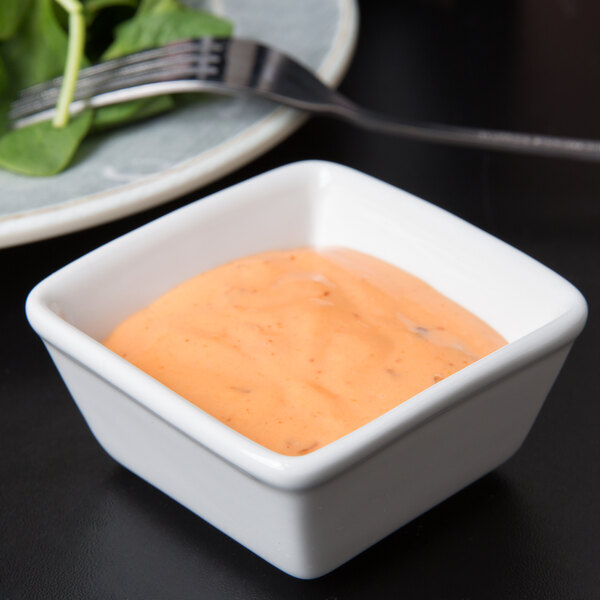 A white square Tuxton TuxTrendz ramekin filled with orange sauce on a table with a salad.